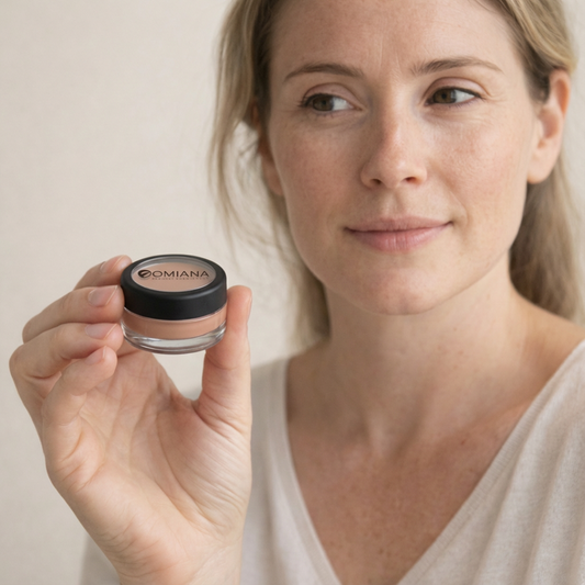 Woman holding a jar of Omiana concealer cream against a plain background